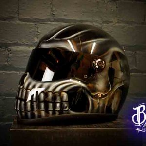 Dark bronze skull motorcycle helmet with metallic airbrushed death head design, detailed teeth and eye sockets against brick wall